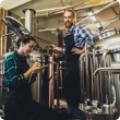 Women with clipboard working at brewery. Brewery worker opening a lid of a beer cask at a craft beer brewery.